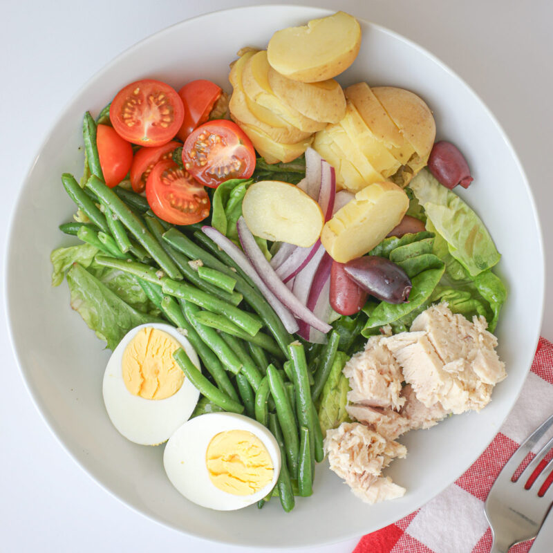 salad nicoise in a white bowl next to a red check cloth with a fork.