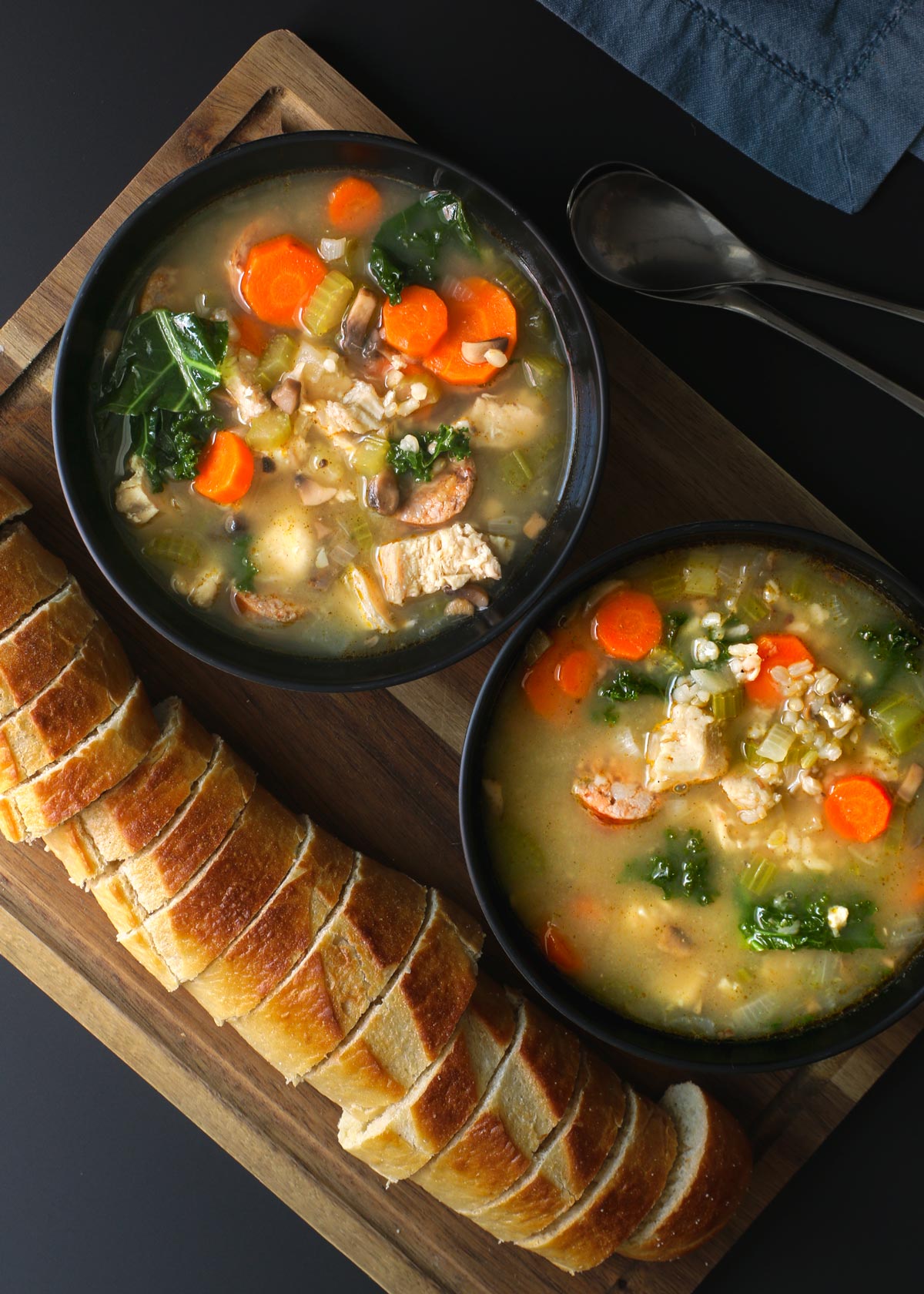 two bowls of turkey and rice soup on a board with a sliced baguette, spoons and napkin nearby.