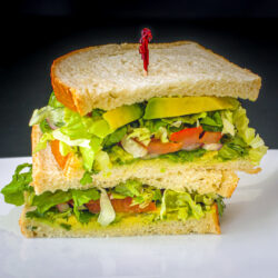 two halves of vegan sandwich with avocado stacked on a plate with a red toothpick in the top half.